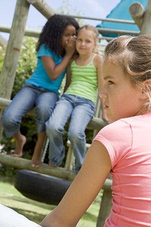 girl alone on playground while other girls whisper together.