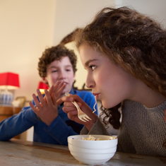 A girl eating with her brother in the background.